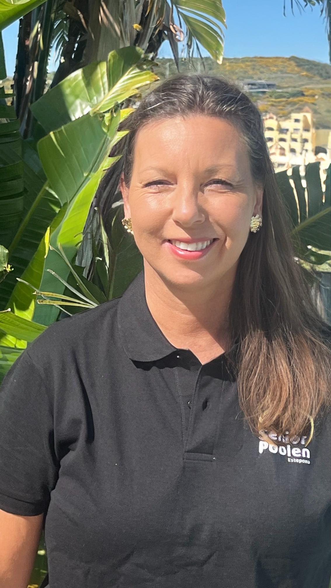 A woman with shoulder-length brown hair, wearing a white blouse, smiles at the camera in a bright room with turquoise and white decorative items on shelves behind her.